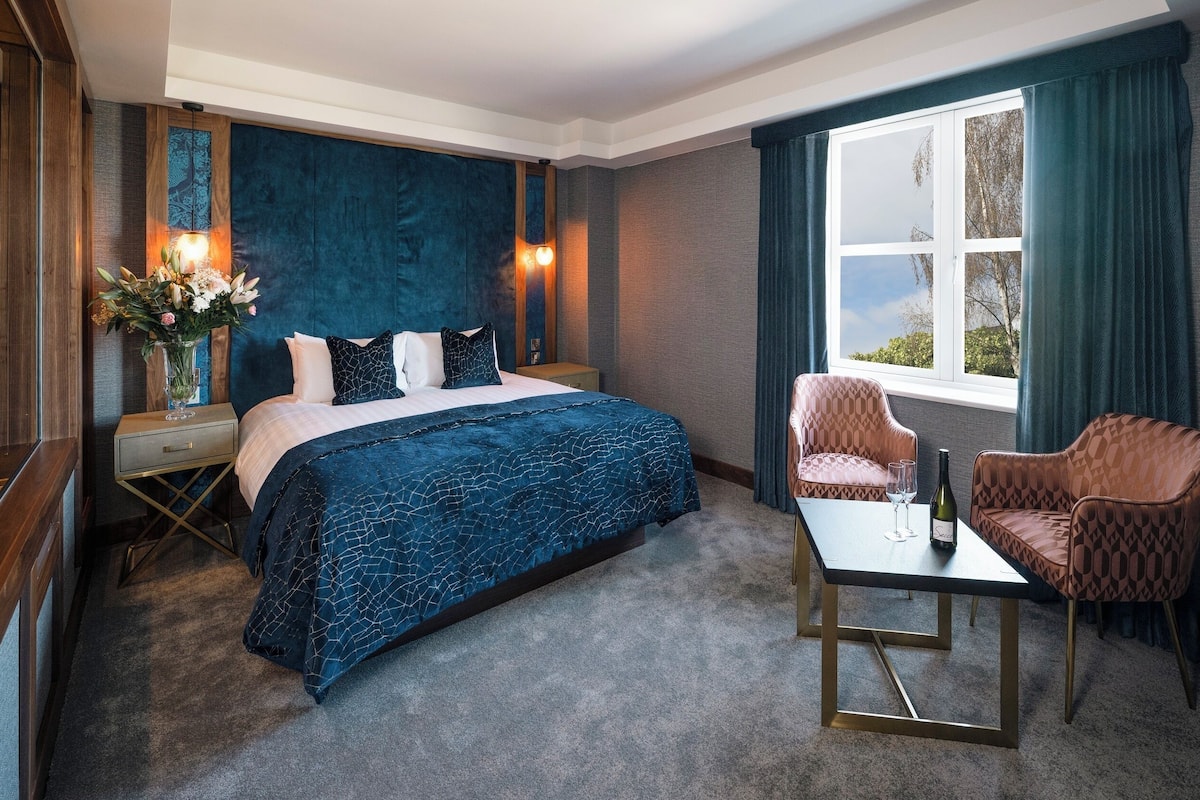 Een hotelkamer met een groot bed, een bureau met stoel, een raam met gordijnen en een vaas met bloemen.