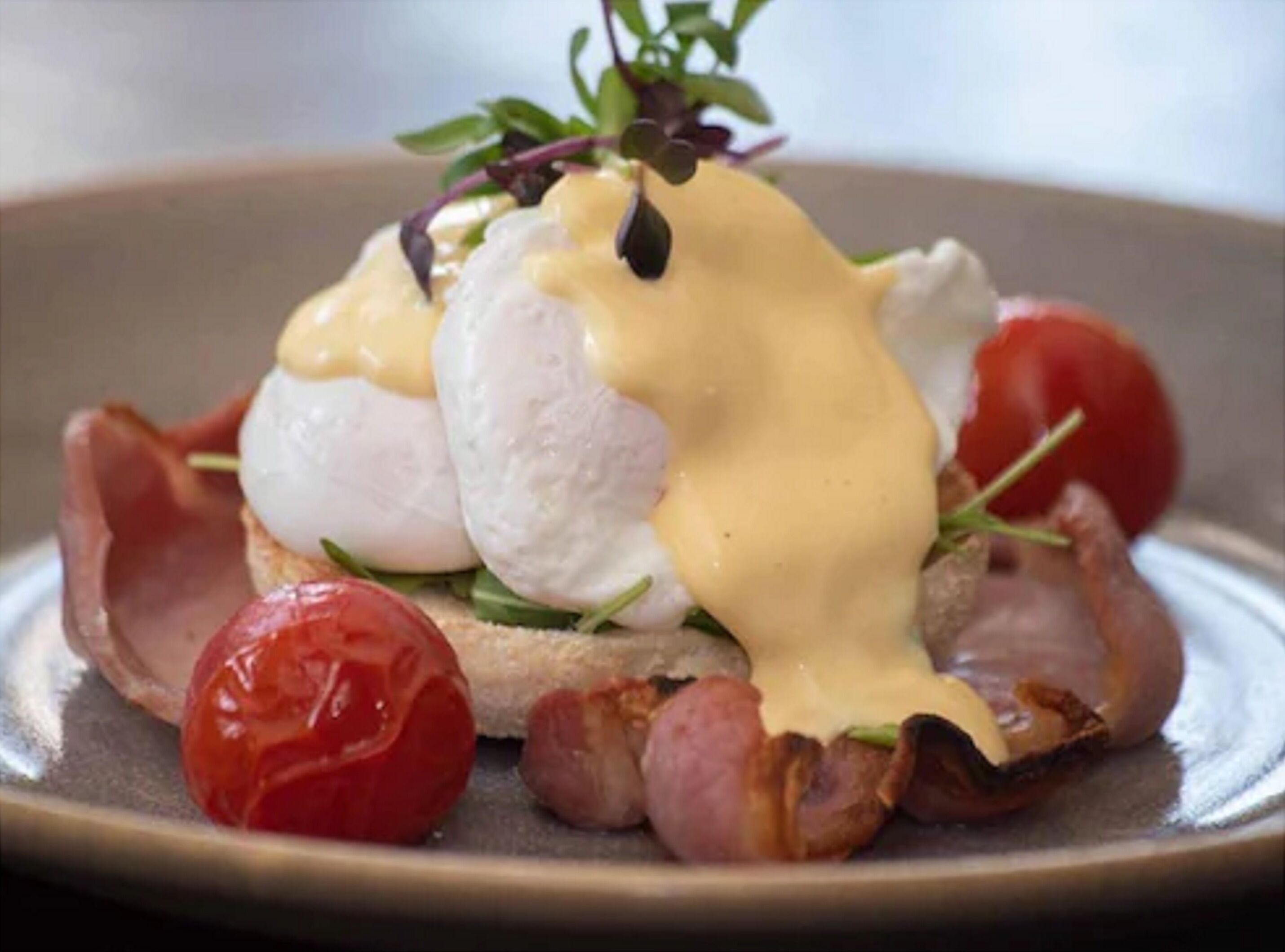 Un plato con huevos pochados, salsa holandesa, tocino y tomates cherry.