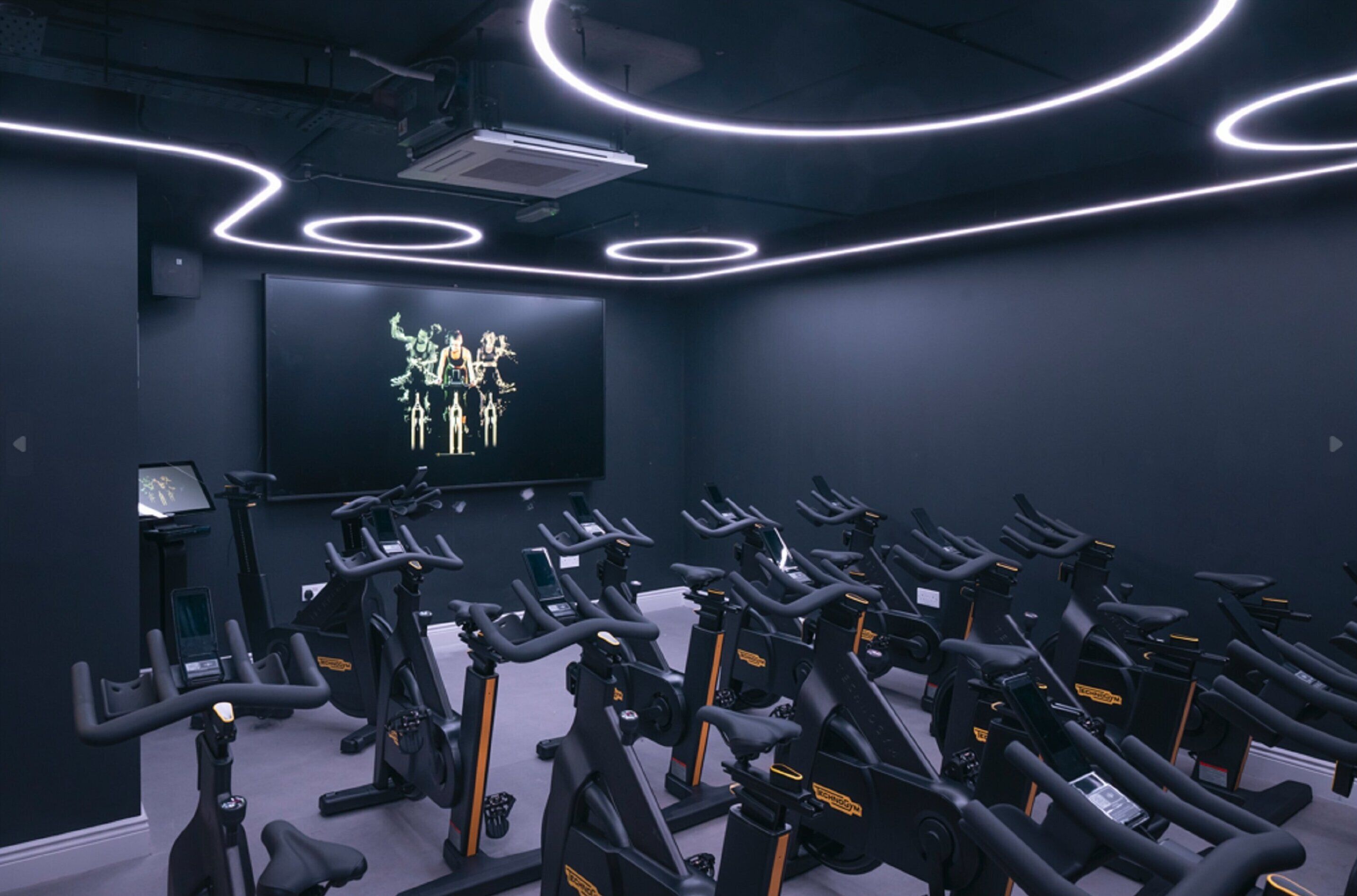 Un gimnasio con varias bicicletas estáticas dispuestas en filas, una pantalla grande mostrando un gráfico y un escritorio pequeño con un ordenador.