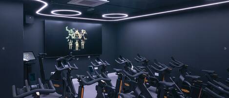 Un gimnasio con varias bicicletas estáticas dispuestas en filas, una pantalla grande mostrando un gráfico y un escritorio pequeño con un ordenador.