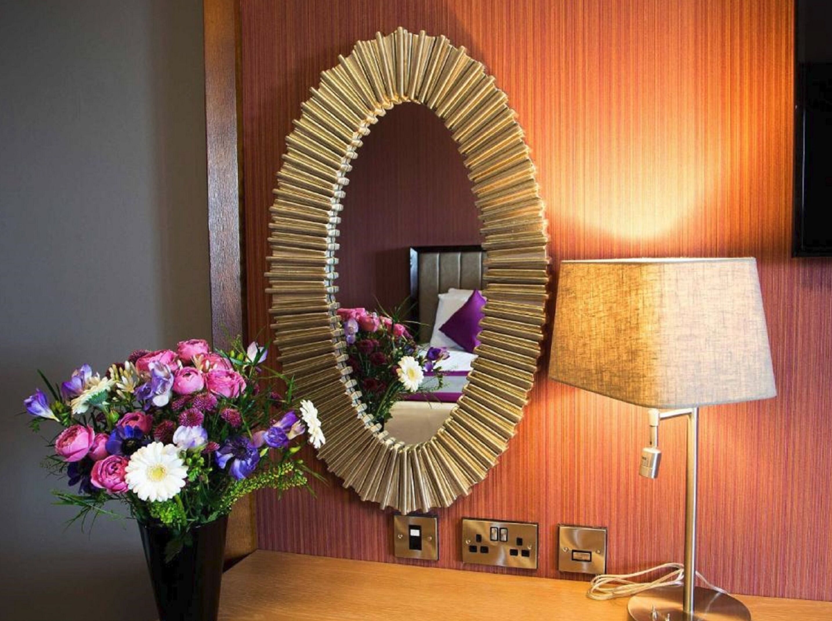 A hotel room with a large mirror, a bedside lamp, and a floral arrangement.