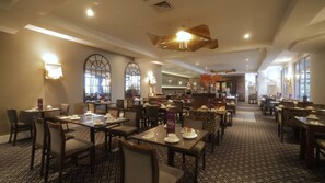 A hotel dining area with multiple tables and chairs, a patterned carpet, and a modern chandelier.