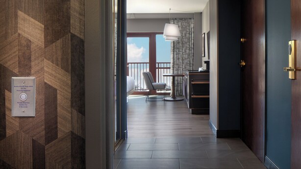 Un couloir d’hôtel avec un mur orné de motifs, une porte dotée d’une poignée en laiton, un bureau avec une lampe et une vue sur un balcon avec une chaise.