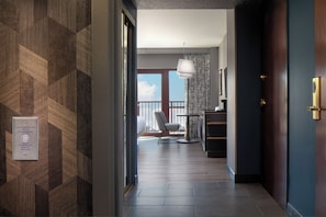 A hotel hallway with a patterned wall, a door with a brass handle, a desk with a lamp, and a view to a balcony with a chair.