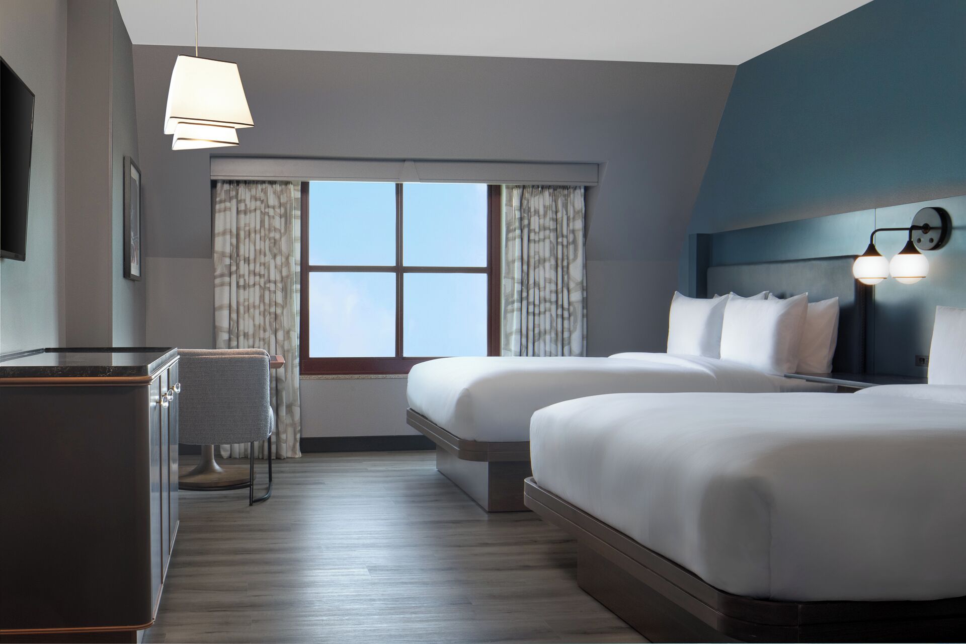 Une chambre d’hôtel avec deux lits, un bureau, une chaise, une grande fenêtre avec des rideaux et une lampe fixée au mur.