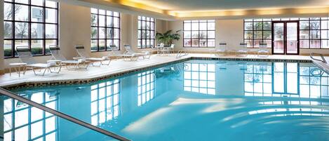 An indoor swimming pool with clear water, sunlit by large windows, and equipped with lounge chairs and a small table.