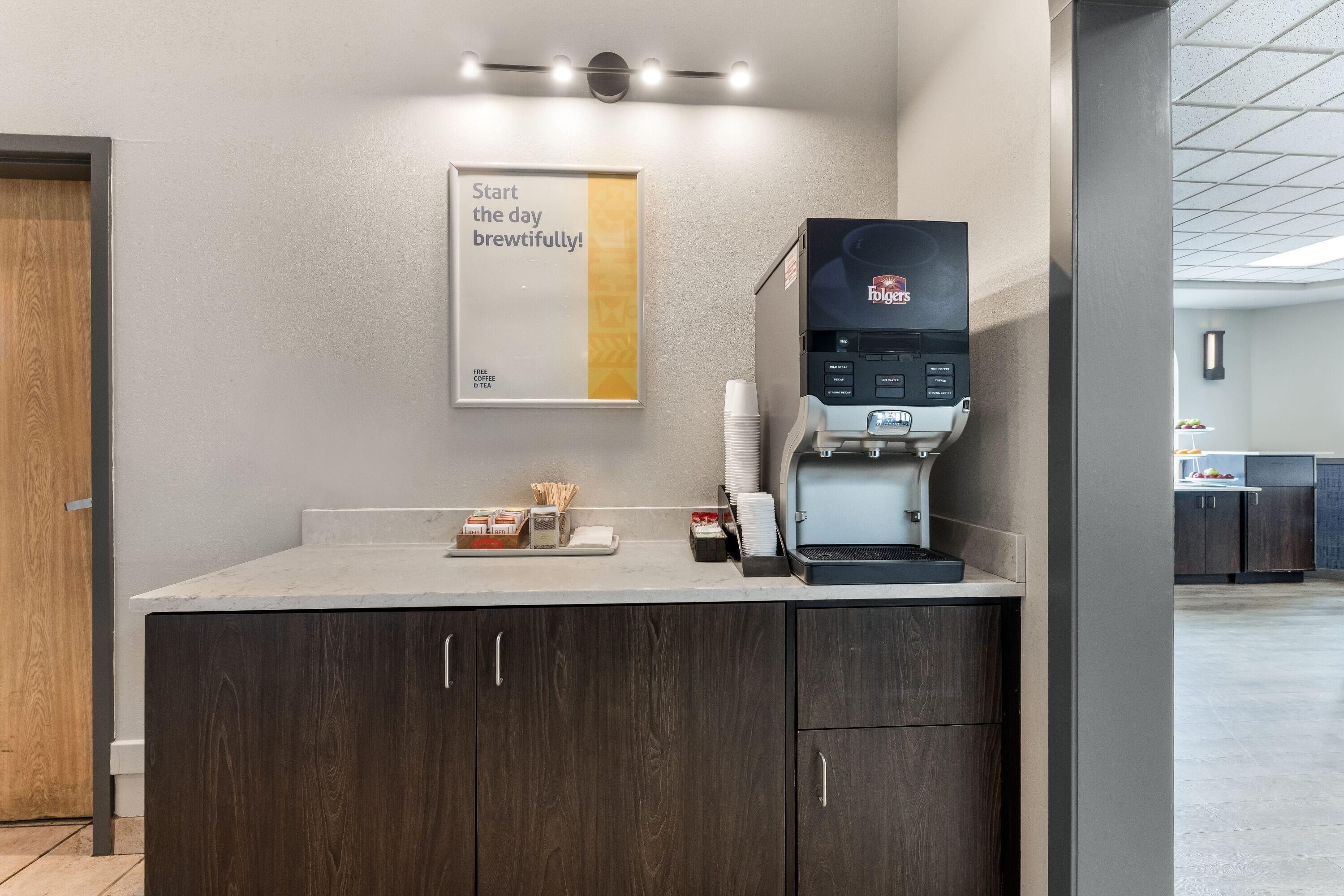 A coffee station with a Folgers coffee maker, cups, and a sign that says 'Start the day brewfully!'