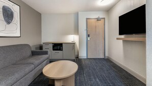 A compact hotel room with a gray sofa, a small table, a kitchenette, a door, and a mounted TV. - Days Inn & Suites by Wyndham Thunder Bay (Thunder Bay)