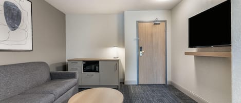 A compact hotel room with a gray sofa, a small table, a kitchenette, a door, and a mounted TV.