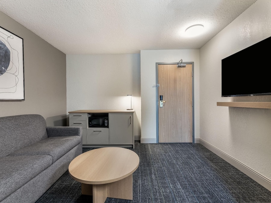 A compact hotel room with a gray sofa, a small table, a kitchenette, a door, and a mounted TV.
