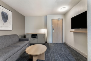 Habitación de hotel compacta con sofá gris, una mesita, kitchenette, una puerta y televisor montado.