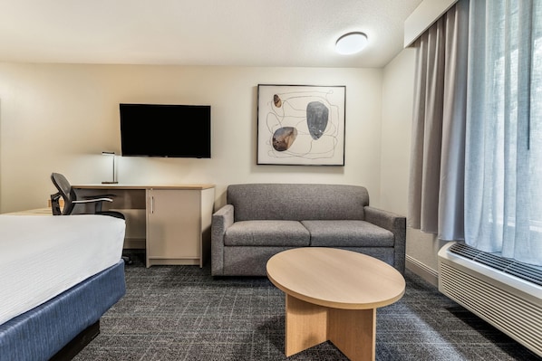 Une chambre d’hôtel comprenant un lit, un bureau, un canapé, une petite table ronde, un téléviseur à écran plat et une fenêtre avec des rideaux.