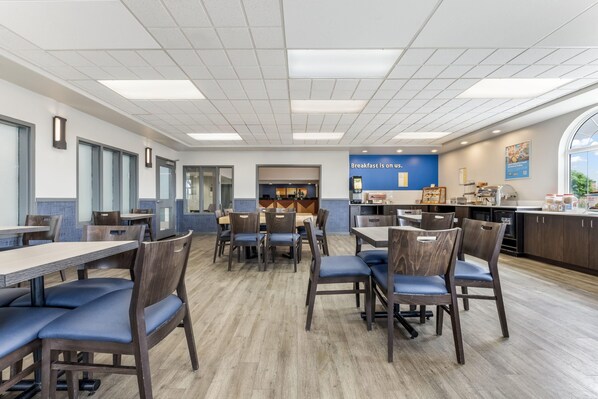 Un espace repas moderne avec des tables et des chaises, une cuisine en arrière-plan et un panneau indiquant le service du petit-déjeuner.