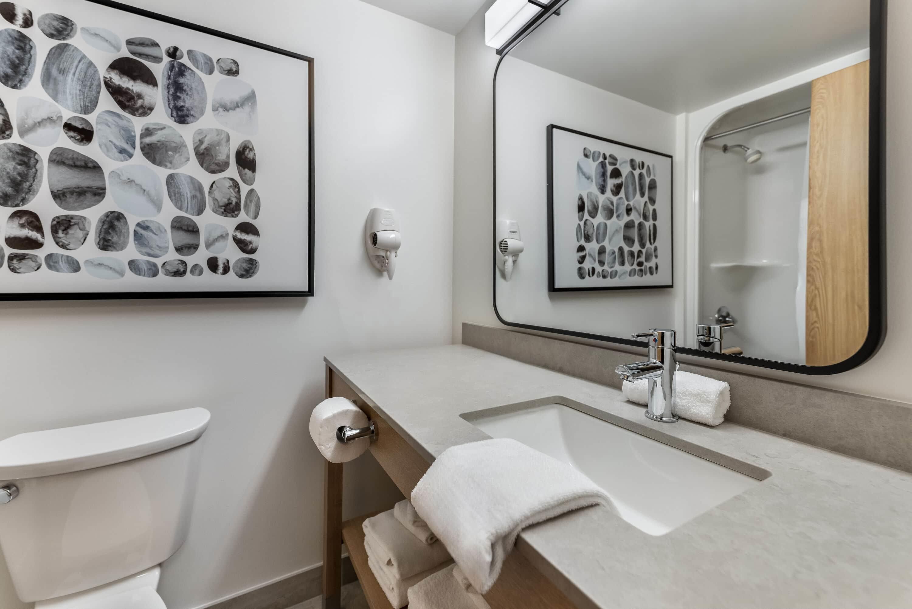 A modern bathroom with a toilet, sink, and mirror. There are framed pictures on the wall and a towel rack.