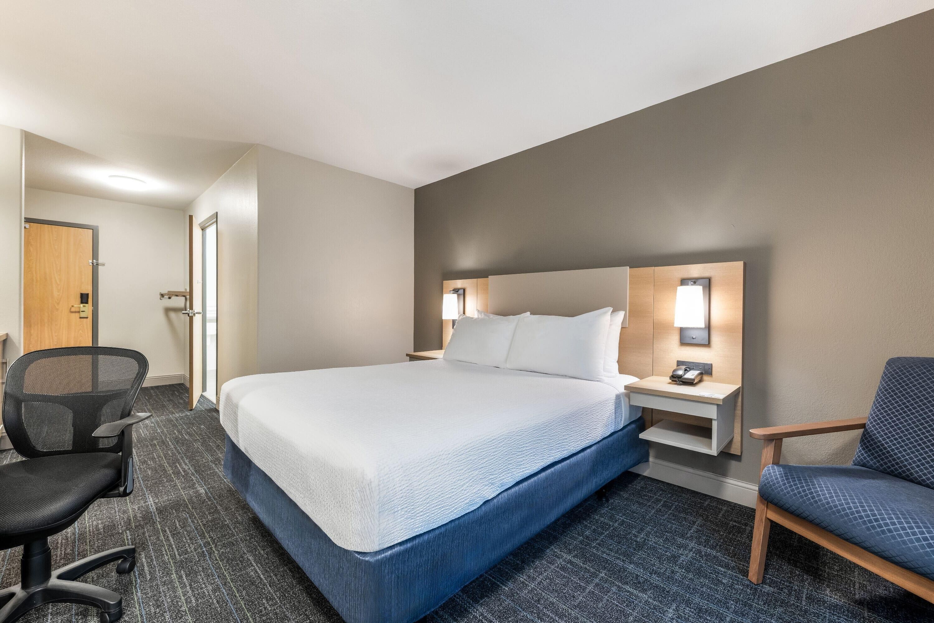 A hotel room with a bed, a desk, a chair, and a nightstand with a lamp.