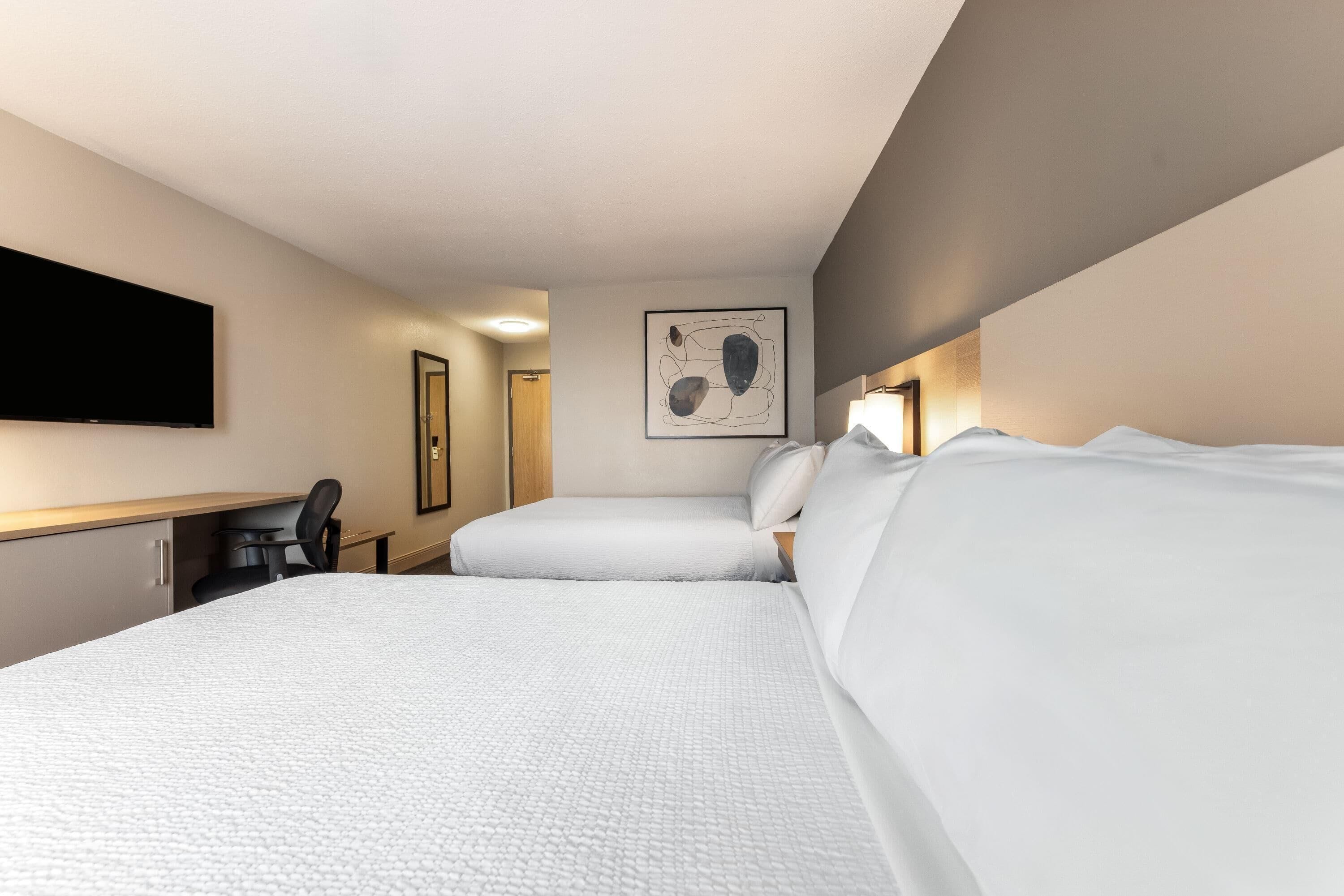 A hotel room with a bed, a desk, a chair, a flat-screen TV, and a framed artwork on the wall.