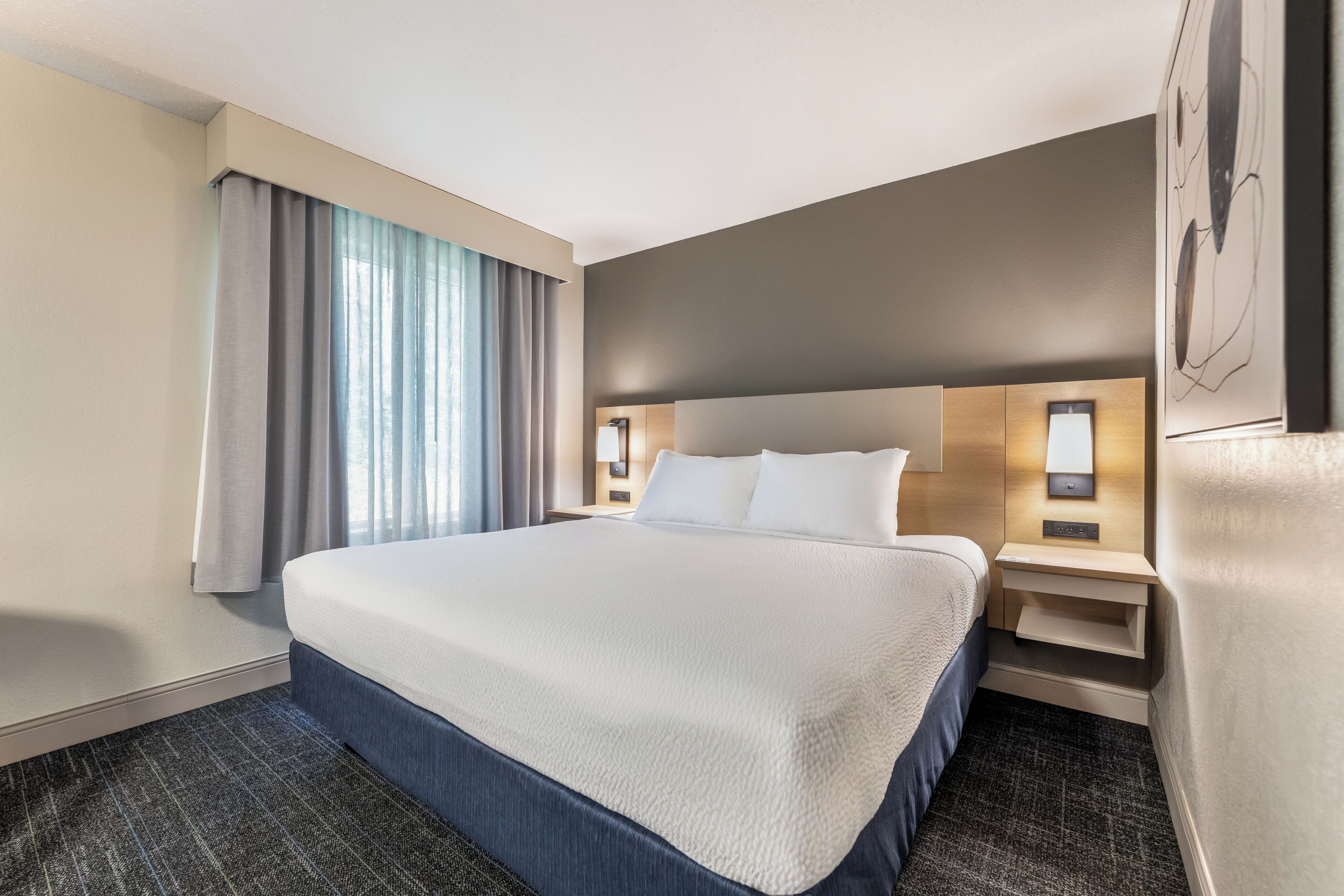 A hotel room with a large bed, two bedside tables, a nightstand, a wall-mounted lamp, and a window with curtains.