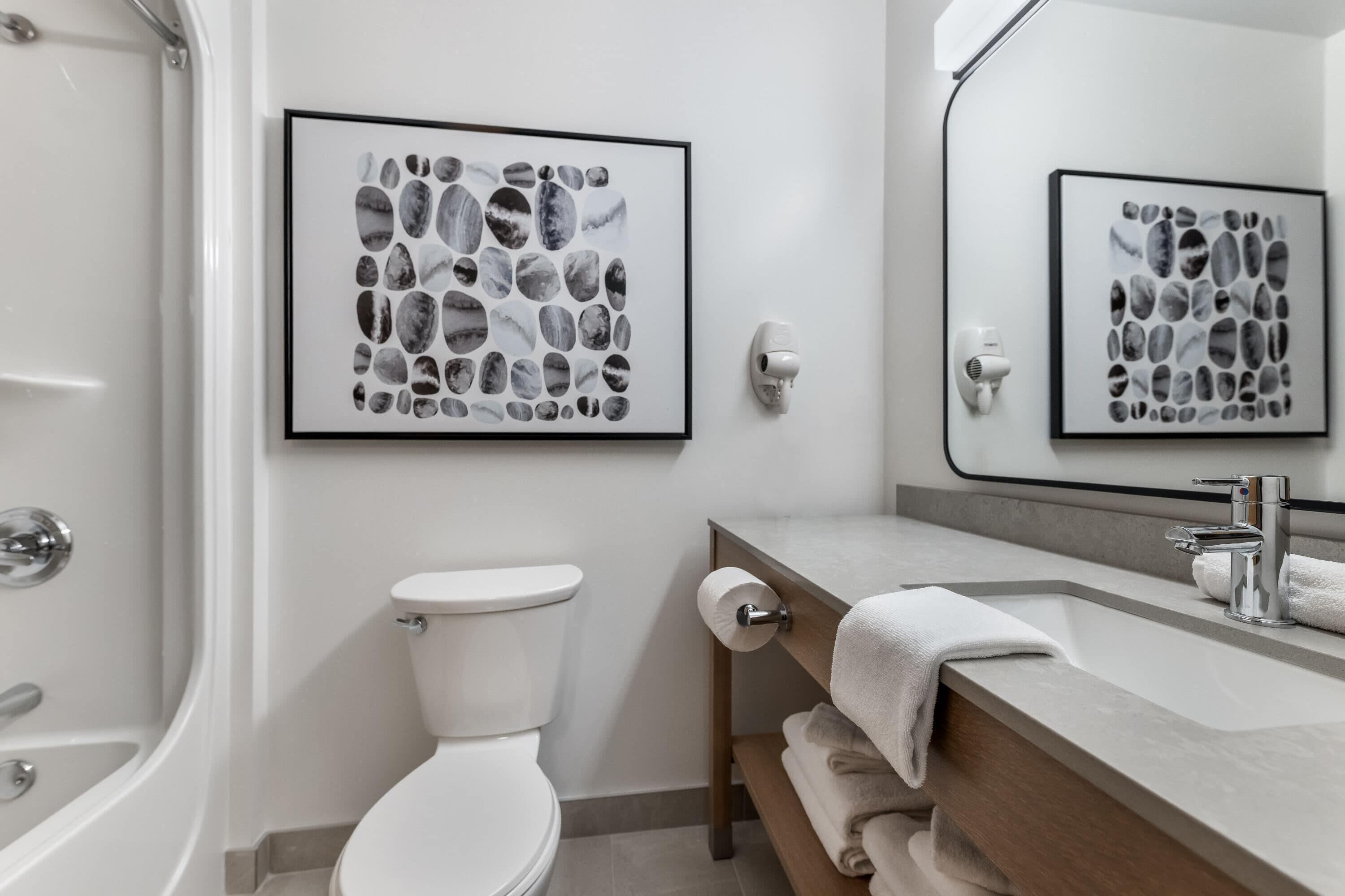 Un baño moderno con inodoro, lavamanos y ducha. Hay obras de arte enmarcadas en la pared y un perchero con toallas.