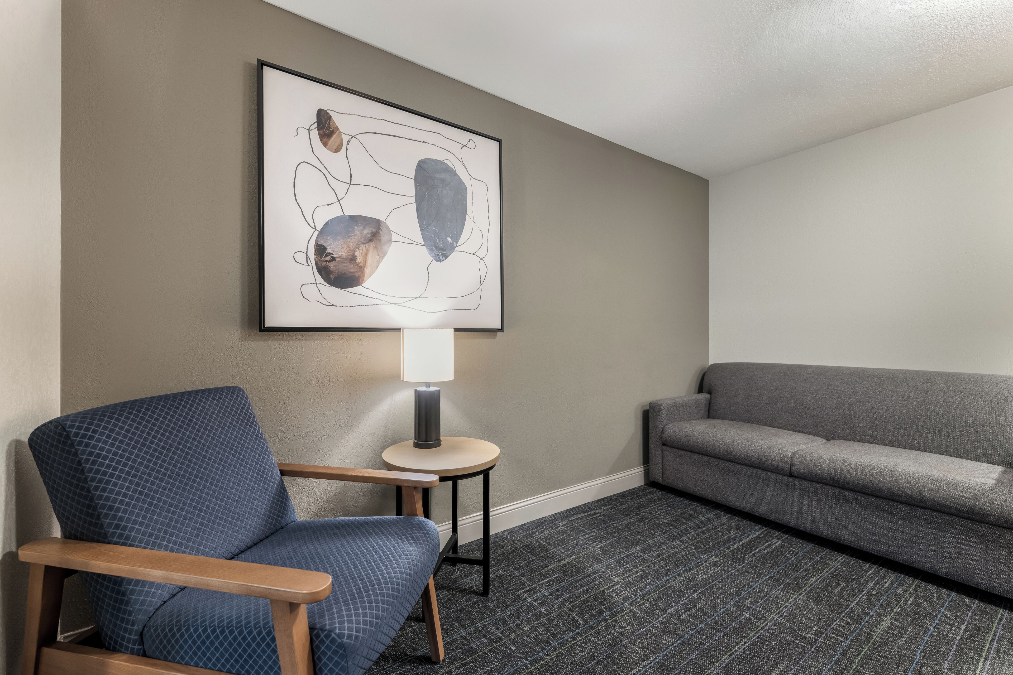 A room with a blue armchair, a grey sofa, a small round table with a lamp, and a framed abstract artwork on the wall.
