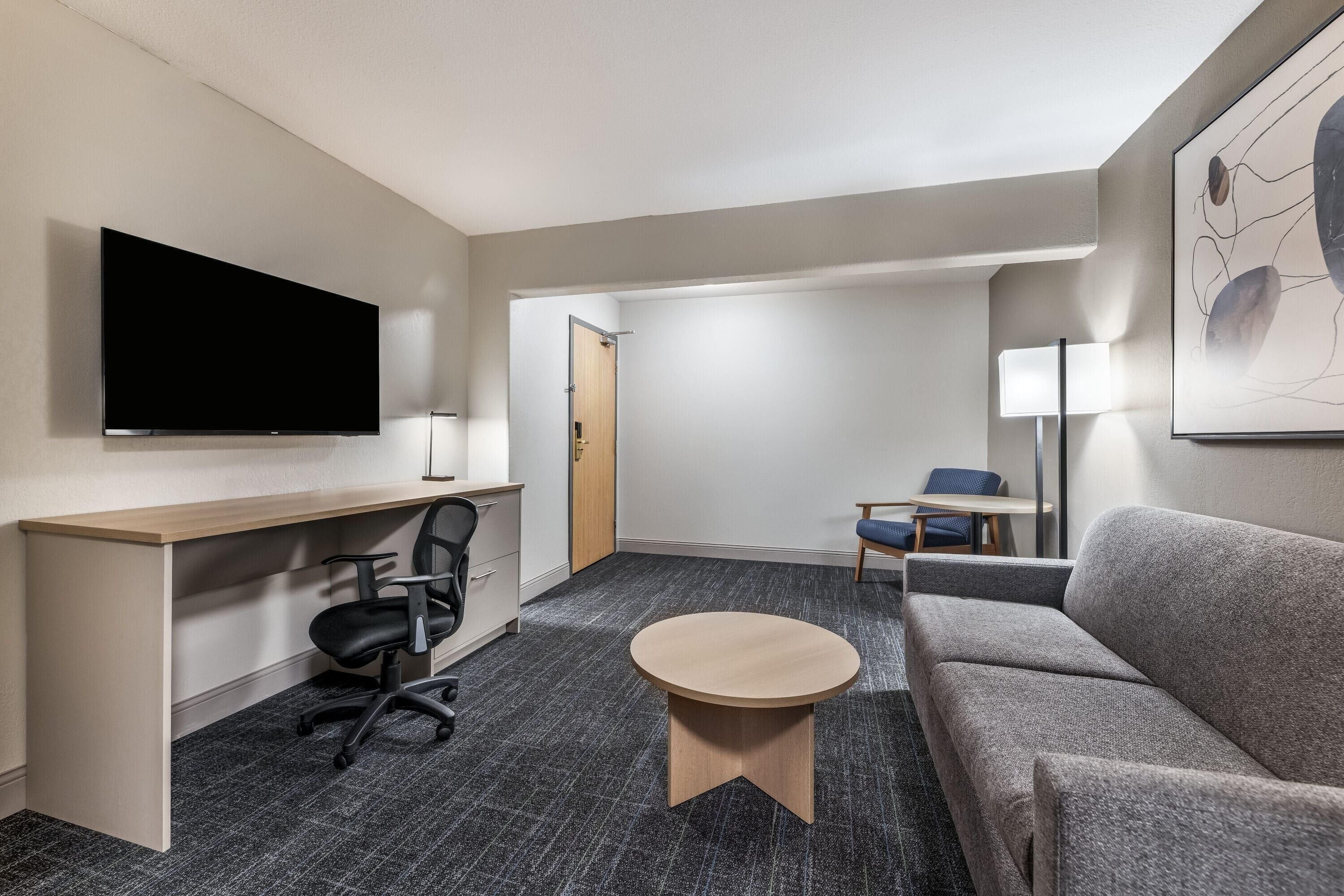 A hotel room with a flat-screen TV, a desk, a chair, a sofa, a small round table, and a lamp.