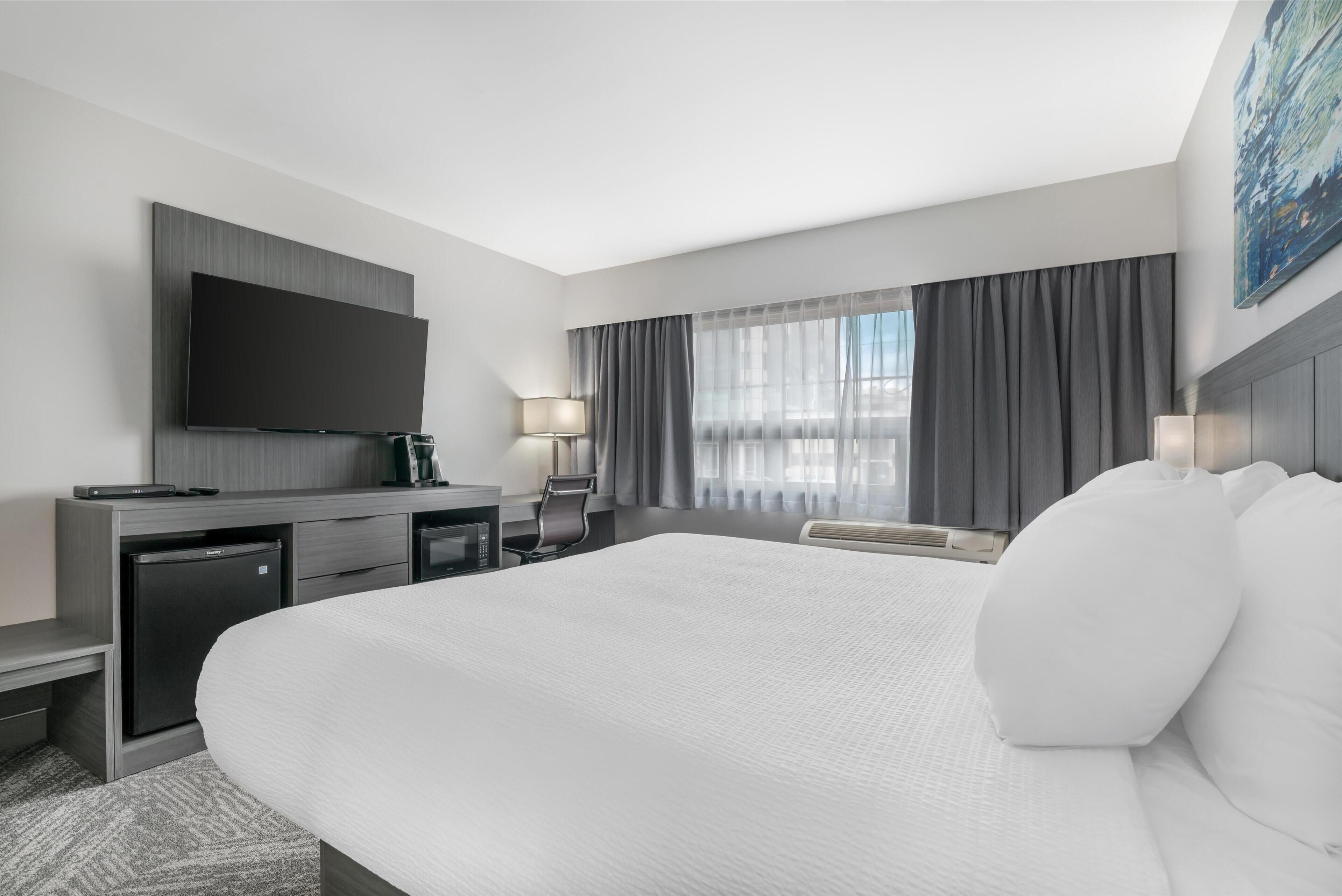 Ein modernes Hotelzimmer mit einem großen Bett, einem flachbildfernseher an der Wand, einem Schreibtisch mit Computer, einer Minibar, einem Sessel und einem Fenster mit Vorhängen.