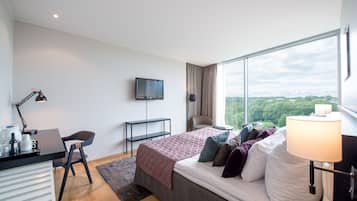 A modern hotel room with a bed, desk, chair, TV, and large windows offering a city view.
