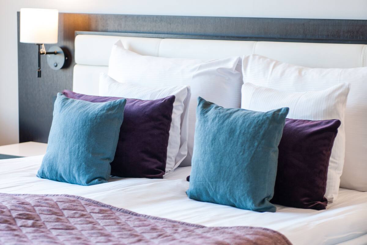 Un letto rifatto con cura, coperta trapuntata, due cuscini in tonalità di blu e viola, e una testiera con una lampada.