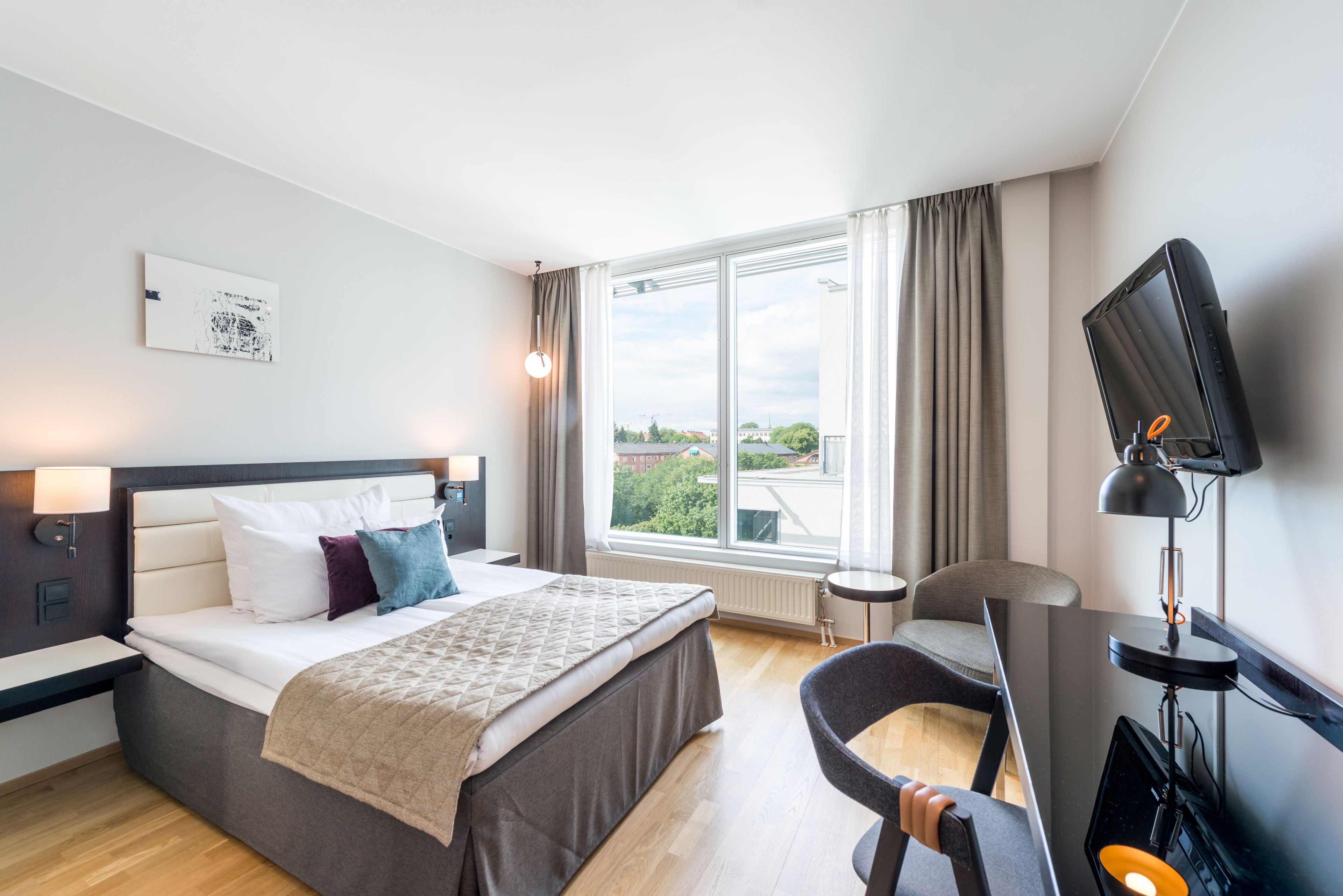 Ett modernt hotellrum med en stor säng, ett skrivbord, en stol, en TV och ett fönster med utsikt över byggnader och träd.