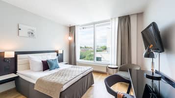 Ett modernt hotellrum med en stor säng, ett skrivbord, en stol, en TV och ett fönster med utsikt över byggnader och träd.