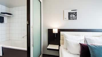 Quarto de hotel com banheira, cama com almofadas brancas e coloridas, luminária na parede e uma moldura com imagem.