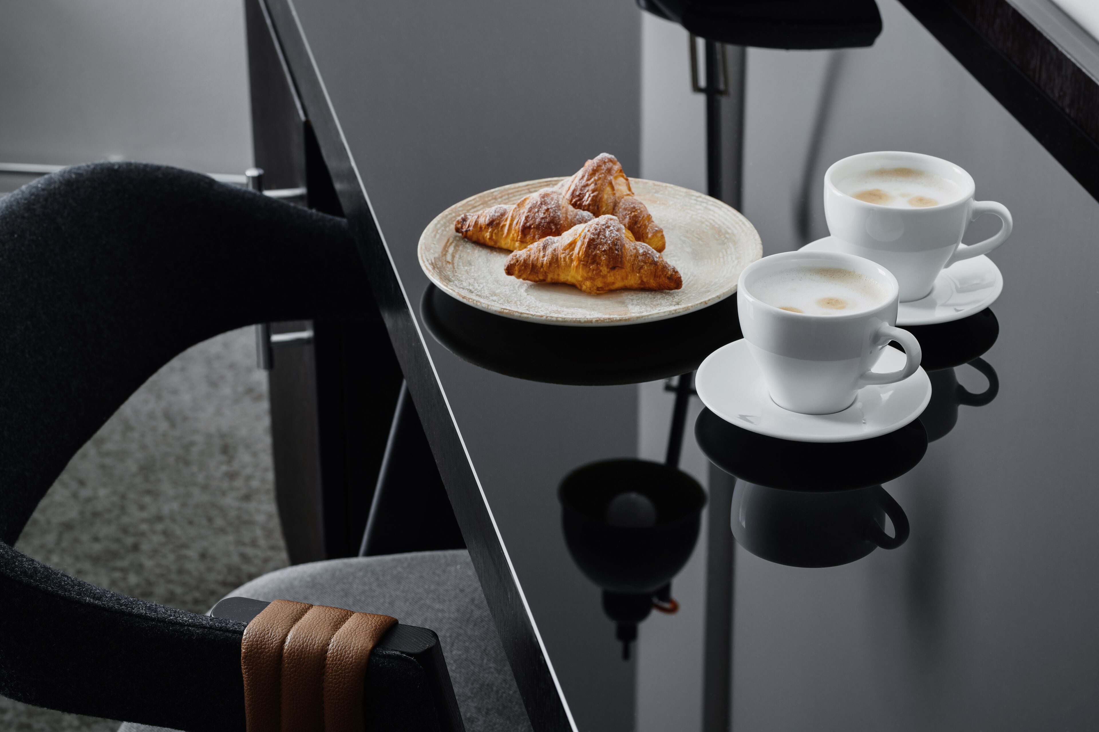Ein Tisch mit Croissants und zwei Tassen Kaffee, deren Spiegelung auf einer glänzenden Oberfläche zu sehen ist.