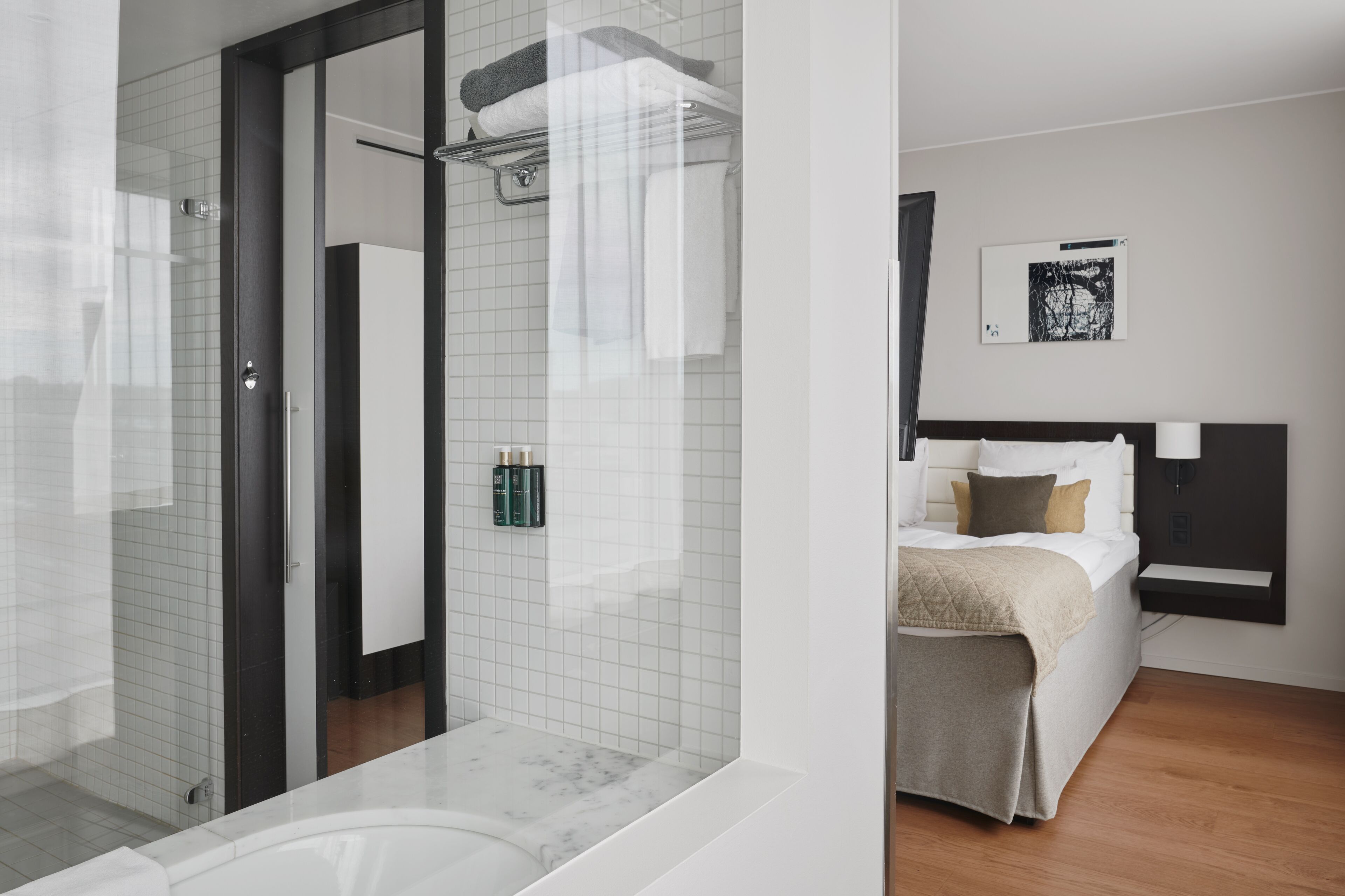 A modern hotel room with a glass shower enclosure, a bed with a headboard, and a small bedside table.