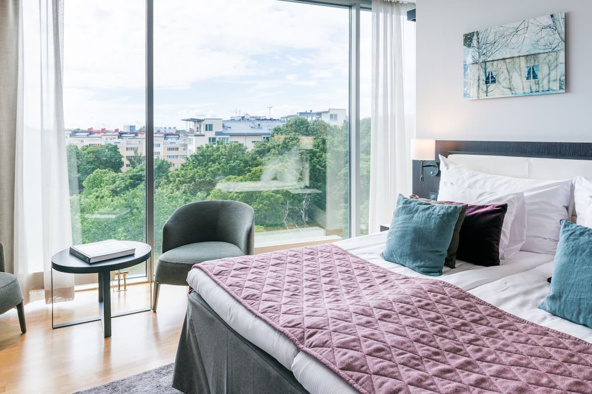 Una moderna camera d'albergo con un ampio letto, vista sulla città, un piccolo tavolo e una sedia.