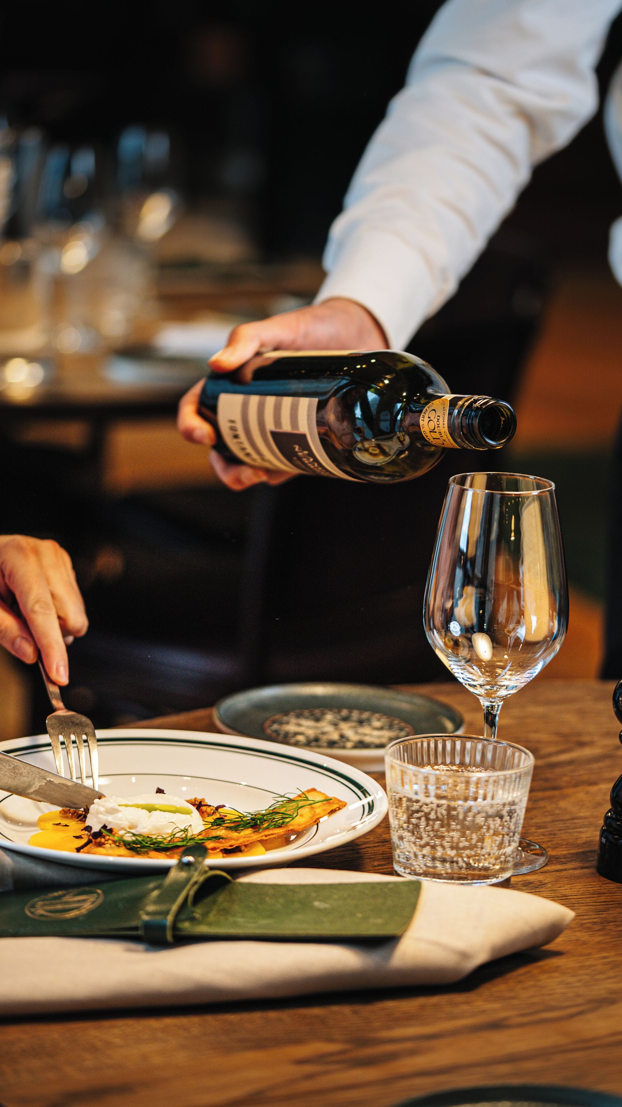 Une personne verse une boisson dans un verre sur une table à manger, où se trouvent des assiettes de nourriture et une fourchette.