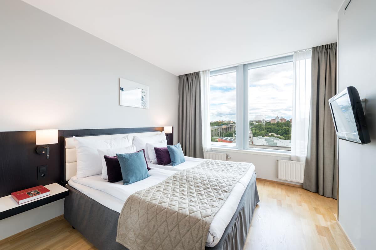 Una camera d'albergo moderna con un letto grande, un comodino con un libro, vista sulla città attraverso ampie finestre e un televisore a schermo piatto montato a parete.