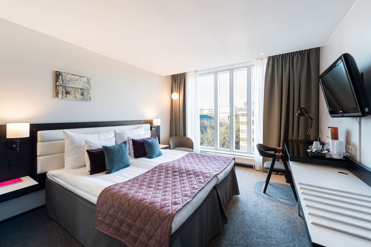 Una camera d'albergo con un letto grande, una scrivania con televisione a schermo piatto, una sedia e una finestra con tende.