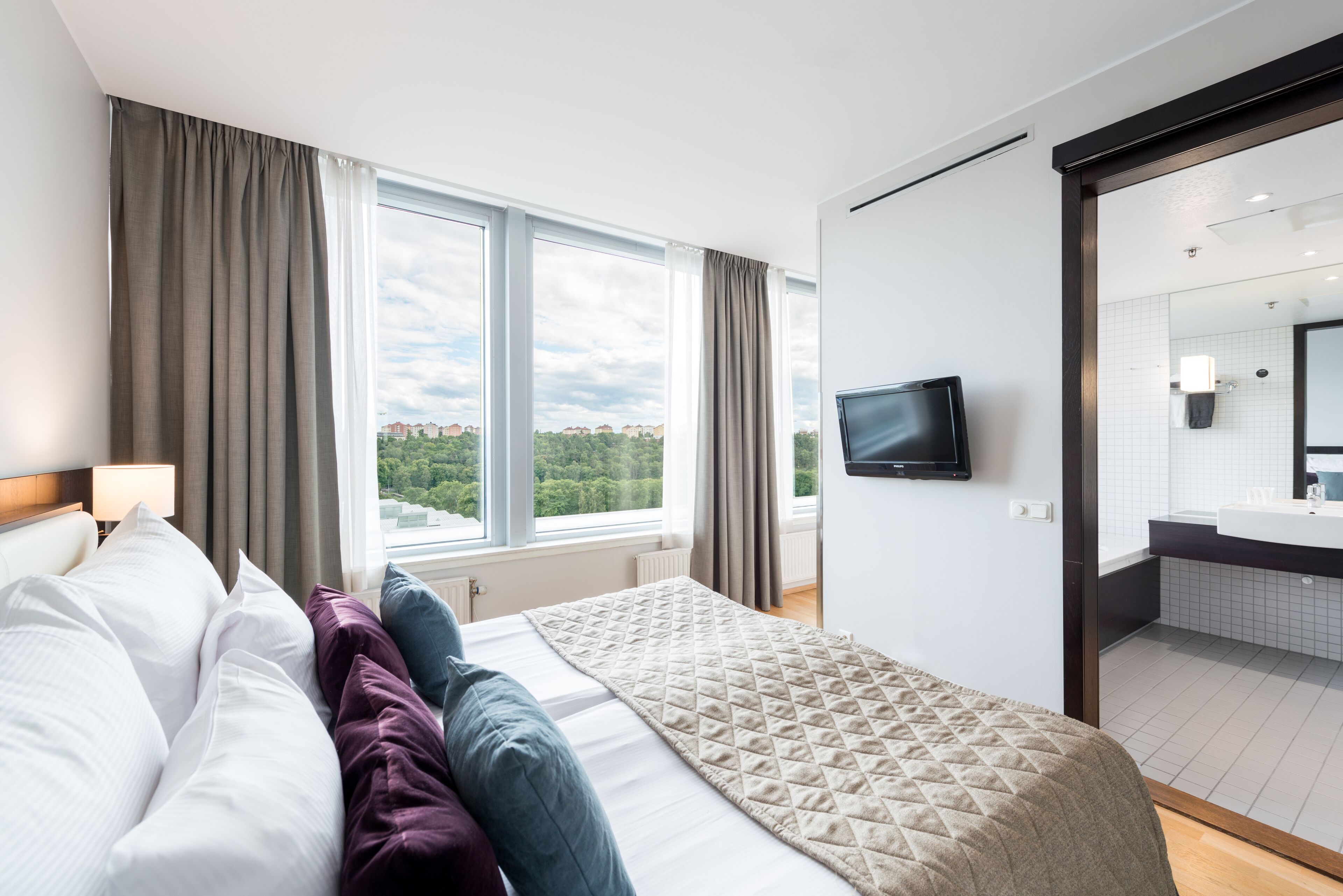 Ein Hotelzimmer mit einem großen Bett, einem an der Wand befestigten Fernseher, Blick auf die Stadt durch große Fenster und einem durch eine offene Tür einsehbaren Badezimmer.