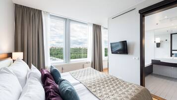 Habitación de hotel con una cama grande, un televisor montado en la pared, vista de la ciudad a través de ventanales amplios y un baño visible a través de una puerta abierta.