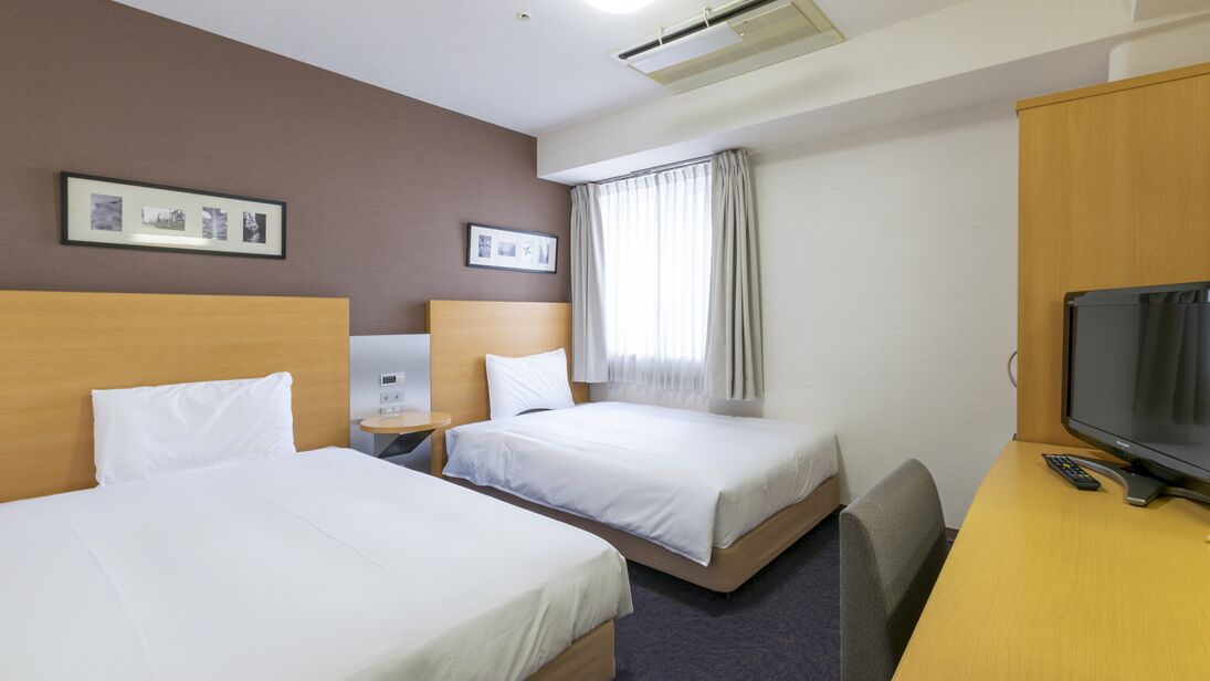 Ein Hotelzimmer mit zwei Betten, einem Schreibtisch mit Fernseher, einem Sessel, einem Fenster mit Vorhängen und gerahmten Bildern an der Wand.
