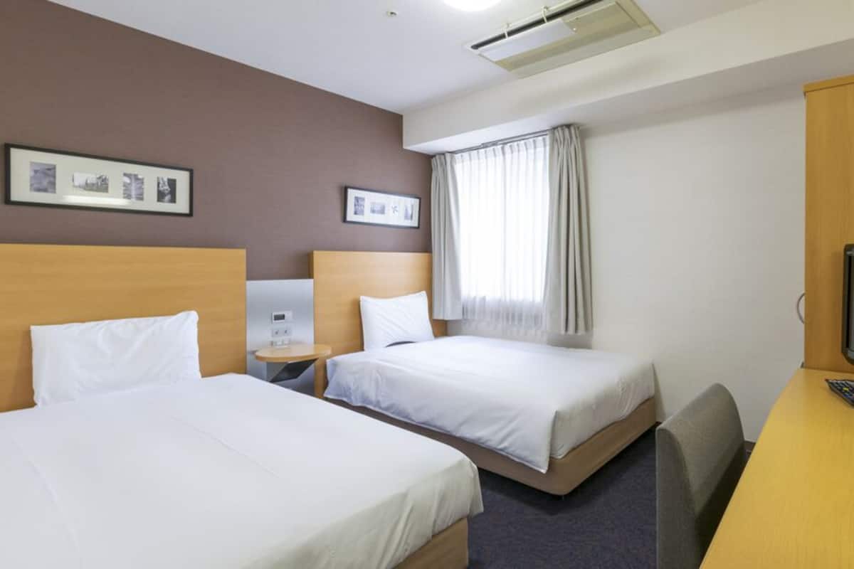 Een hotelkamer met twee bedden, een bureau met een televisie, een stoel, een raam met gordijnen en ingelijste foto's aan de muur.