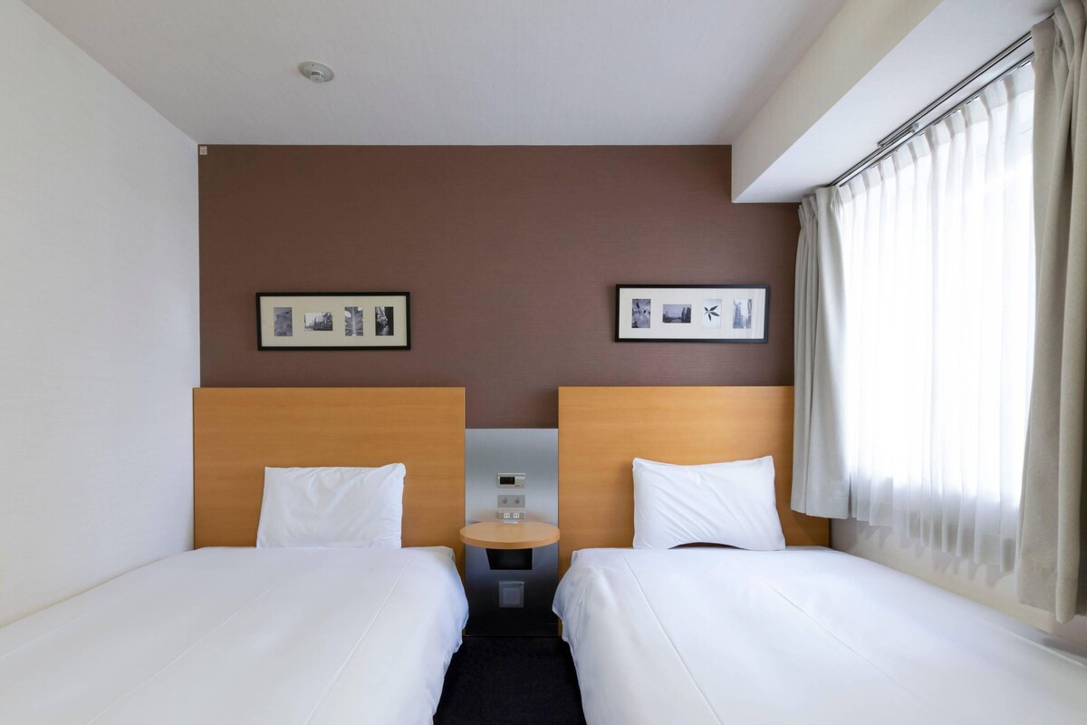 Habitación de hotel con dos camas individuales, una mesita de noche con reloj digital y dos cuadros en la pared.