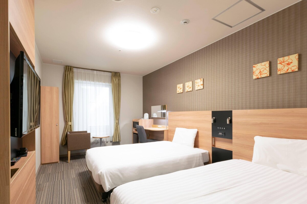 Een hotelkamer met twee bedden, een bureau, een stoel, een televisie en een raam met gordijnen.