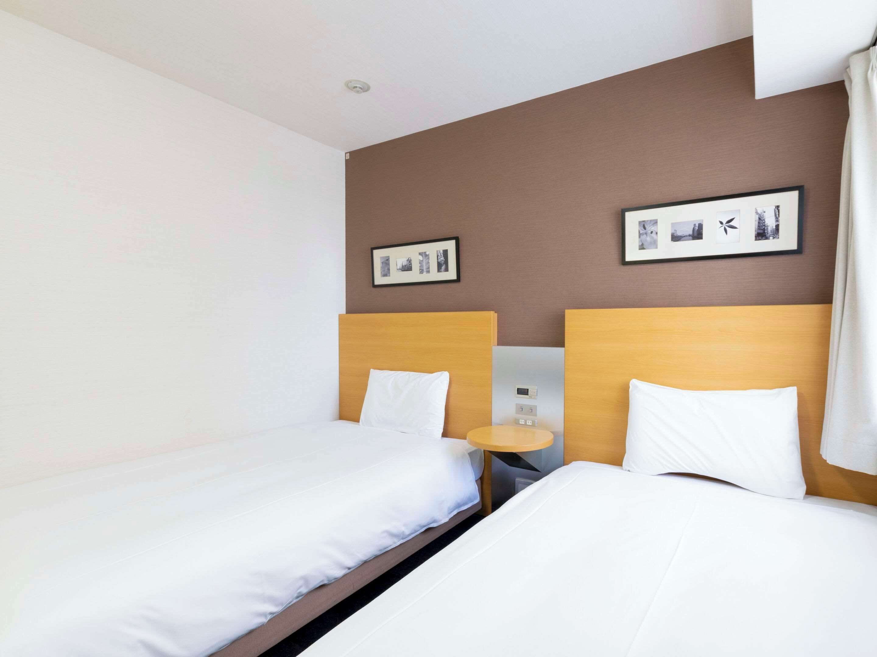 A hotel room with two single beds, a small table, and a wall-mounted control panel.