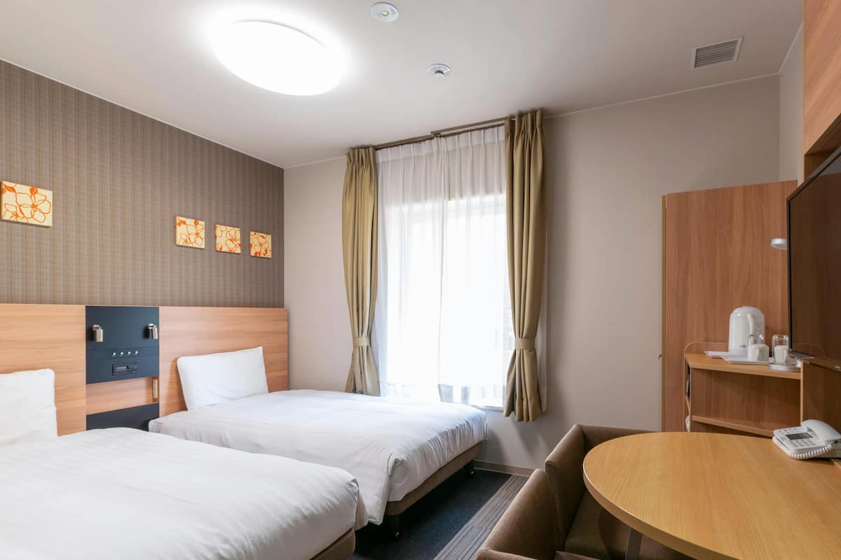 Een hotelkamer met twee bedden, een ronde tafel, een bureau met telefoon, en een raam met gordijnen.
