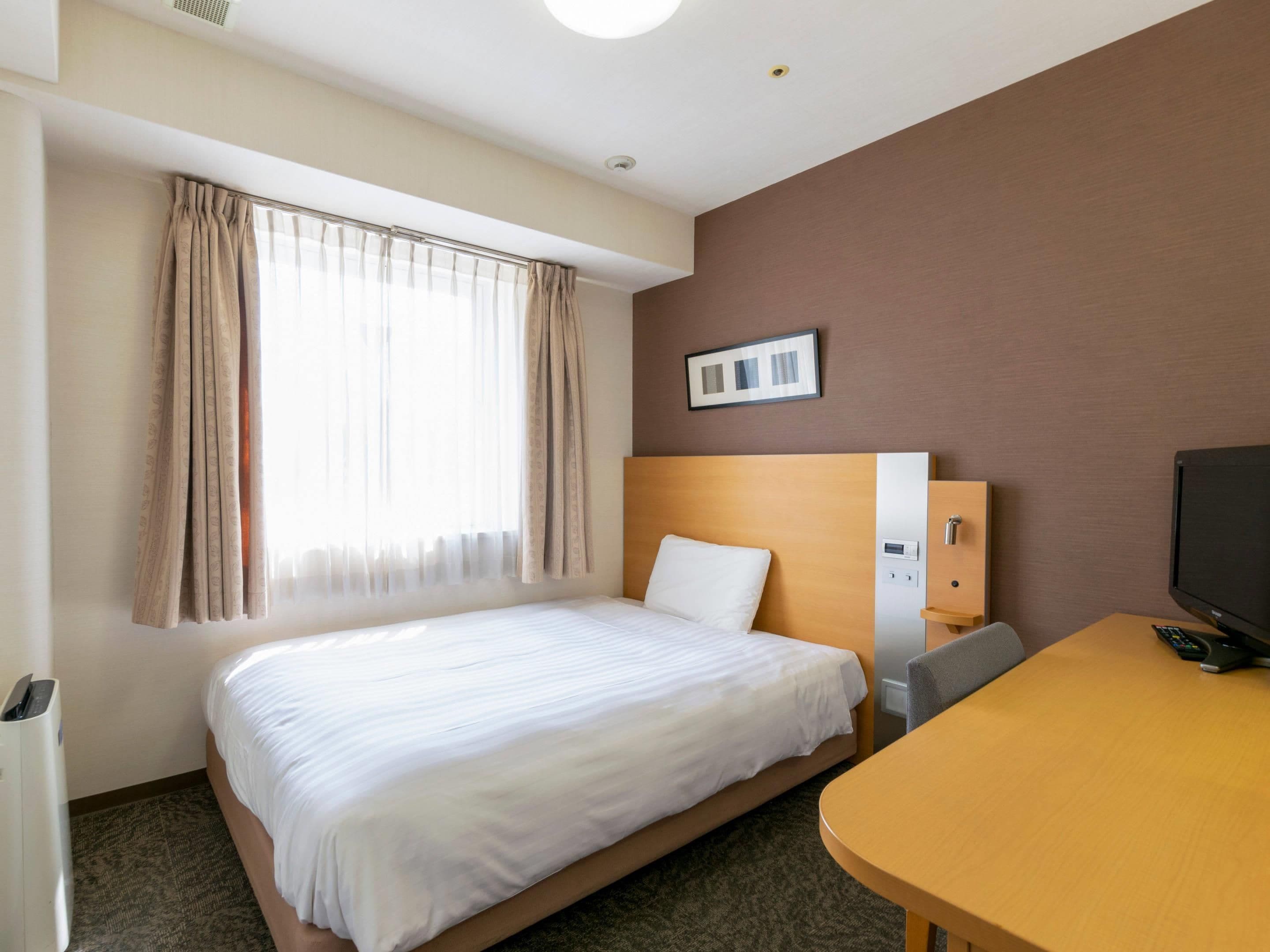 A hotel room with a single bed, a desk, a television, and a window with curtains.