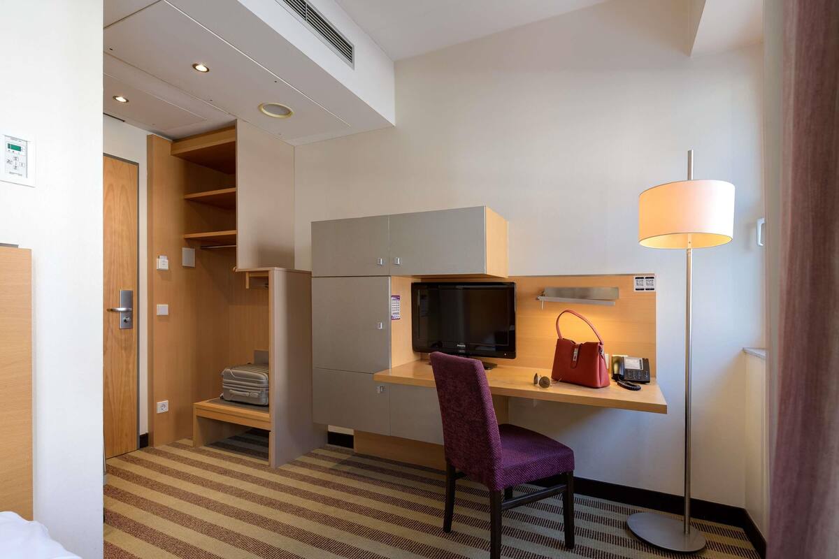 A hotel room with a bed, a desk with a computer, a chair, a TV, a lamp, and a wardrobe.