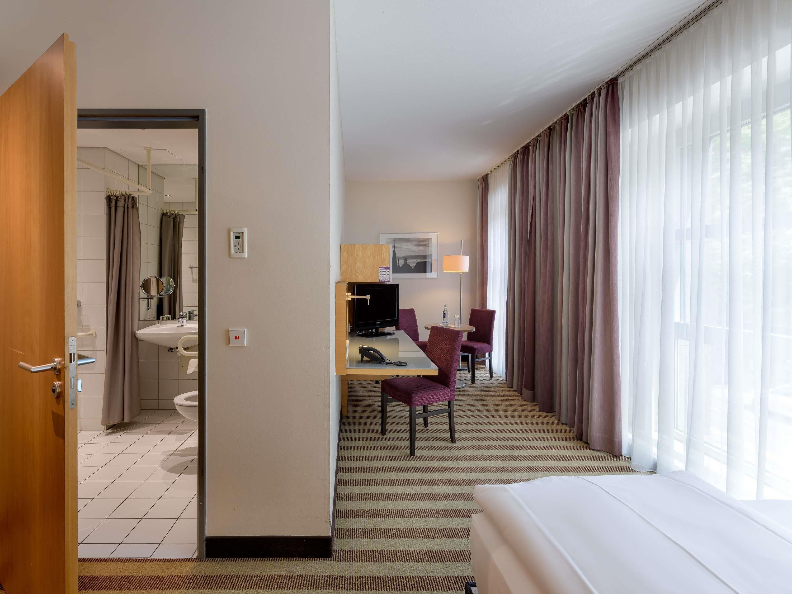 A hotel room with a bed, a desk with a chair, a TV, a window with curtains, a bathroom with a sink and mirror, and a door.