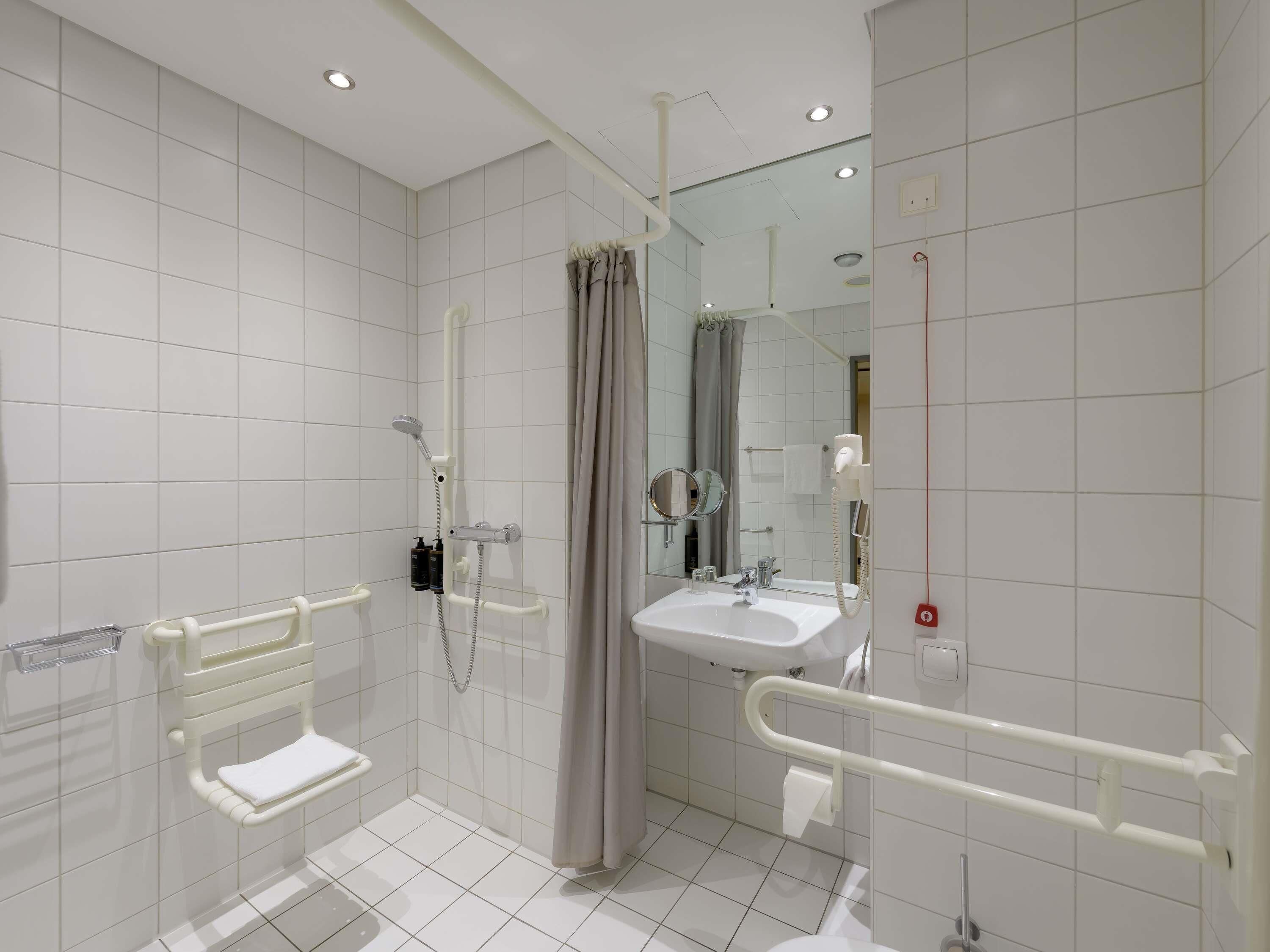 A modern bathroom with white tiles, a shower with a handheld showerhead, a sink with a mirror, and a toilet.