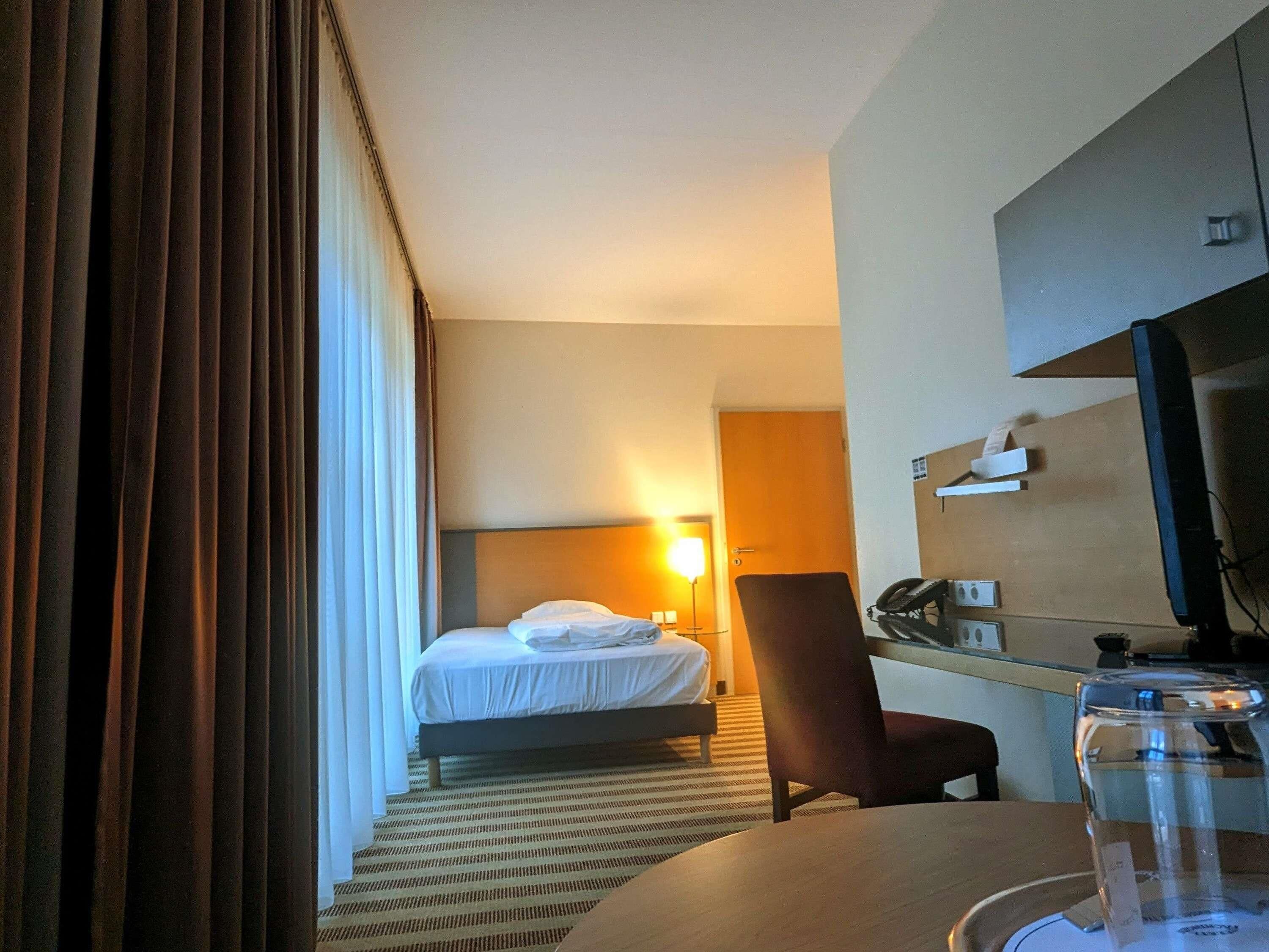 A hotel room with a bed, a desk, a chair, a television, and a glass of water on the table.