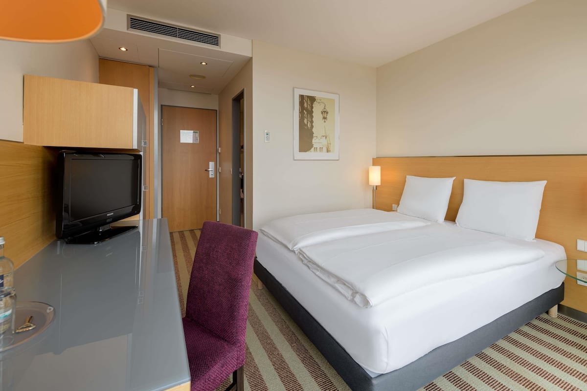 A hotel room with a bed, a desk with a television, a chair, and a wall-mounted lamp.
